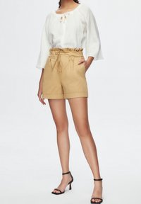Lightweight white blouse with a round neckline and three-quarter sleeves. Tan shorts with elastic waistband and rolled cuffs. Black strappy heels.