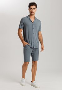 Pánská sada krátkého rukávu košile na knoflíky a šortky z jemné, světle šedé látky. Vlastnosti zahrnují límec, přední knoflíky a boční kapsy. Oblečeno s bílými botami.