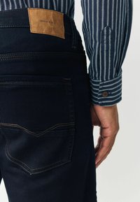 Tmavě modré džíny z denimu s hnědou koženou nášivkou "DENIM SUPPLY." Mají kontrastní prošití a zadní kapsu s jemným vzorem.