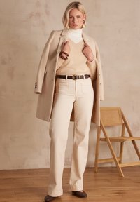 Cappotto di lana beige sopra un dolcevita crema, abbinato a un maglione beige chiaro e pantaloni crema a vita alta. Cintura scura e scarpe marroni.