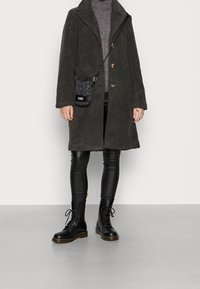 Abrigo de forro polar oscuro con dos botones, cuello de tortuga gris, leggings de cuero sintético negros y botas de combate negras con cordones. Pequeño bolso de estampado en el hombro.