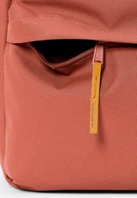 Mochila de tela en color óxido con un bolsillo con cremallera y un acento de cuero marrón claro. Presenta un material texturizado y un tirador de cremallera suave.