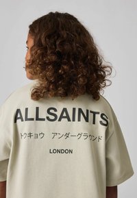 Heledbeez oversize T-särk lühikeste varukatega, mille tagaküljel on suur must tekst: "ALLSAINTS," jaapani tähed ning "LONDON."