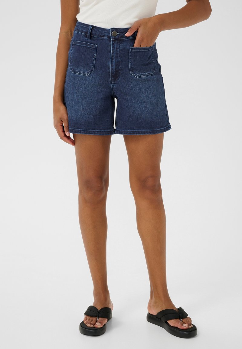 Femme portant un short en jean bleu foncé avec des poches plaquées à l'avant et des sandales noires, debout sur fond blanc.
