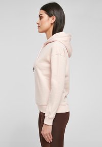 Lys rosa hettegenser med snøring, avslappet passform, ribbestrikkede mansjetter og nederkant. Bært med mørkebrune leggings, vist mot en grå bakgrunn.