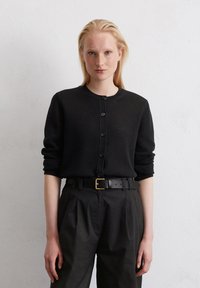 Schwarze Strickjacke mit Rundhalsausschnitt, Knopfverschluss und langen Ärmeln, kombiniert mit hoch taillierten schwarzen Hosen und einem schwarzen Gürtel.