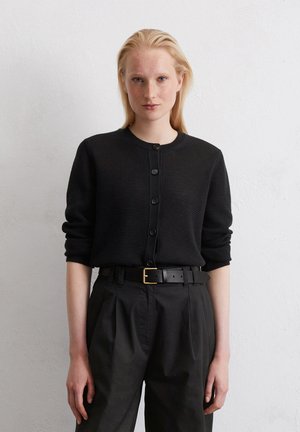 Schwarze Strickjacke mit Rundhalsausschnitt, Knopfverschluss und langen Ärmeln, kombiniert mit hoch taillierten schwarzen Hosen und einem schwarzen Gürtel.