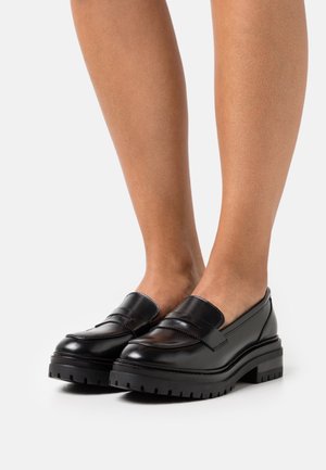 Black leather loafers with a chunky rubber sole, featuring a stitched detail across the vamp and a smooth finish. Classic design, slip-on style.