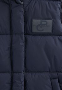 Navyblå vadderad väst i slätt tyg med en rektangulär lapp med "POLO CLUB"-logotyp och horisontell sömnadsdetalj.