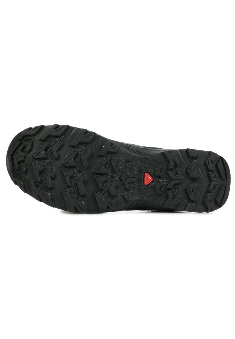 Suela de zapato de goma negra con tapones de tracción multidireccionales, superficie texturizada y un prominente acento triangular rojo cerca del talón.