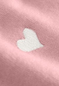 Lichtroze stof met een zachte textuur heeft een witte geborduurde hartvormige detail. Het hart is omrand met een gestructureerde afwerking.