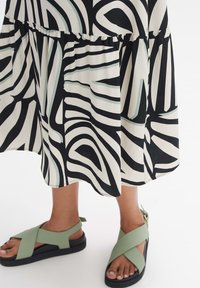 Black and white patterned dress with ruffled hem paired with green cross-strap sandals featuring a black sole and adjustable ankle strap.