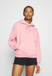 Vaaleanpunainen huppari, jossa on kiristysnauhainen huppu ja etutasku. Pehmeä materiaali ja pieni rintalogo. Yhdistetty mustien shortsien kanssa.