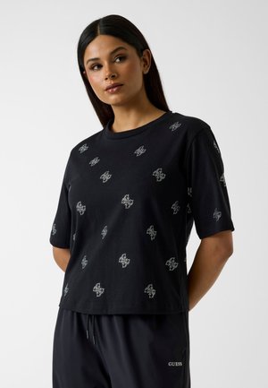 Femme aux longs cheveux foncés portant un t-shirt noir avec de petits motifs papillons blancs et un pantalon noir à cordon.