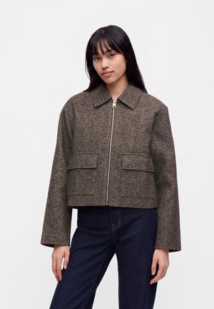 Junge Frau trägt eine braune Wolljacke mit Reißverschluss und großen Fronttaschen sowie dunkelblaue Jeans, steht vor einem schlichten Hintergrund.