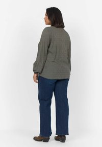 Langärmlige, gemusterte Bluse in Grau mit weißen Punkten, kombiniert mit blauen Jeans. Die Bluse hat einen abgerundeten Saum und leicht gepuffte Ärmel.