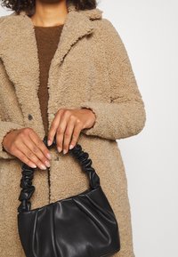 Beige, texturovaný kabát z umělé kožešiny s šálovým límcem; černá kabelka s nařaseným uchem držena v rukou. Hnědý svetr pod kabátem.