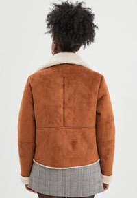 Braune Kunstwildlederjacke mit einem weißen Lammfellkragen und -manschetten, die eine taillierte Form und ein karierter Saum hinten aufweist.