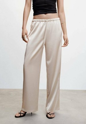 Vrouw draagt beige satijnen broek met wijd uitlopende pijpen en elastische taille, gecombineerd met zwarte sandalen met bandjes en hak, staand op een betonnen vloer.