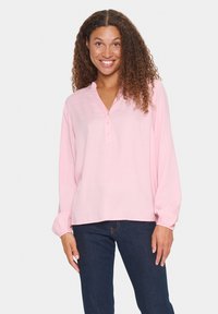 Blusa rosa de manga larga con cuello henley, que presenta tres botones y una tela texturizada. Combinada con jeans de mezclilla oscura.
