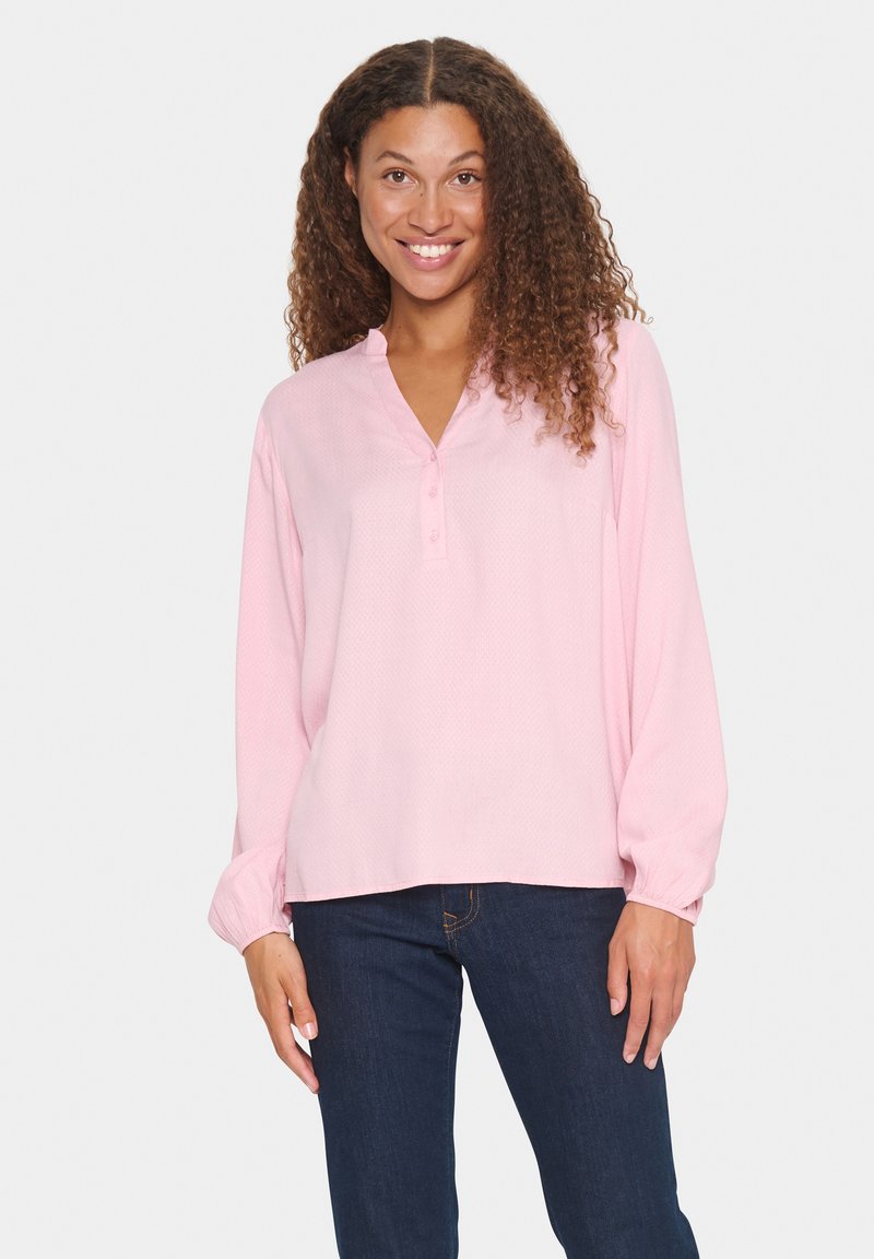 Blusa rosa de manga larga con cuello henley, que presenta tres botones y una tela texturizada. Combinada con jeans de mezclilla oscura.