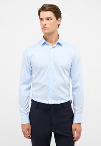 Maglietta azzurra a maniche lunghe con colletto a punte, chiusura a bottoni e maniche con polsini, accoppiata a pantaloni scuri. Texture del tessuto liscia.