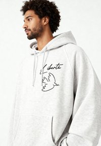 Felpa con cappuccio grigio chiaro con tasca anteriore, presenta una colomba nera ricamata e la parola "Liberté" sopra di essa. Tessuto strutturato con coulisse.