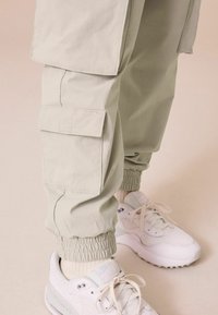 Pantalons cargo beige clair avec plusieurs poches latérales, poignets élastiques, associés à des baskets blanches et des chaussettes beiges sur un fond neutre.