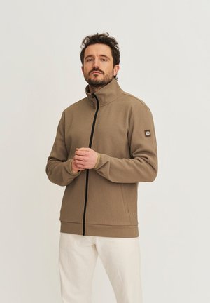 Homme barbu portant une veste marron texturée zippée et un pantalon blanc, debout devant un fond clair uni.
