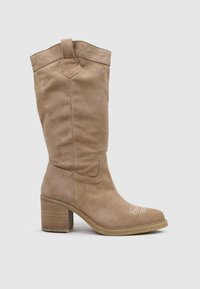 Botín de ante marrón claro con punta cuadrada y tacón cuadrado. Presenta costuras decorativas en la parte superior y una textura suave en toda la bota.