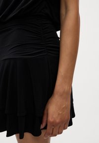 Vestido preto feito de tecido macio, apresentando uma cintura franzida e uma bainha em camadas. Close-up de uma mão descansando contra a saia.