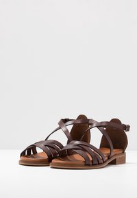 Paire de sandales plates en cuir marron avec plusieurs brides et boucle à la cheville sur fond blanc.