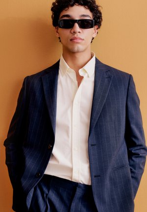 Persoană tânără purtând un costum în dungi negre, o cămașă albă și ochelari de soare negri dreptunghiulari, stând pe un fundal de culoare muștar.