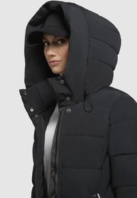 Giubbotto imbottito nero con un grande cappuccio imbottito e dettagli con bottoni a pressione. Presenta una chiusura con zip e una superficie testurizzata.
