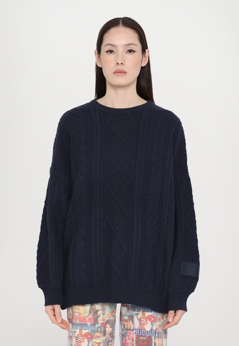 Pull en maille côtelée bleu marine avec un col rond, coupe oversize, poignets côtelés et un motif texturé. Présente une patch logo sur la manche.