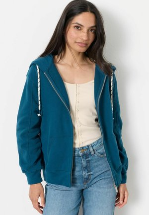 Kvinde med langt mørkt hår og fregner iført en teal lynlåshættejakke over en cremefarvet knapbluse og blå jeans, stående mod en hvid baggrund.