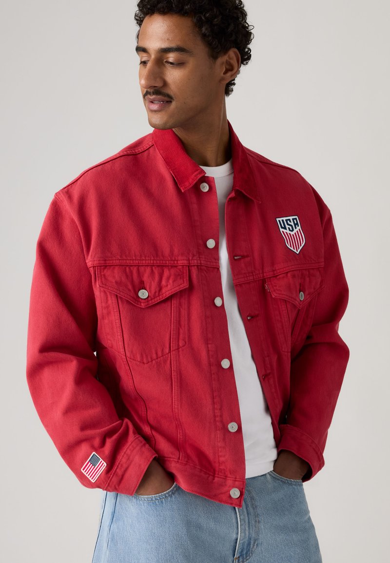 Uomo che indossa una giacca di jeans rossa con una toppa USA sul petto, toppa della bandiera americana sulla manica, camicia bianca e jeans azzurri, mani nelle tasche.