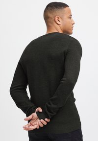 Maglione a maglia testurizzata di colore verde scuro, con scollo rotondo, maniche lunghe e polsini a coste, visto di spalle con le mani unite.
