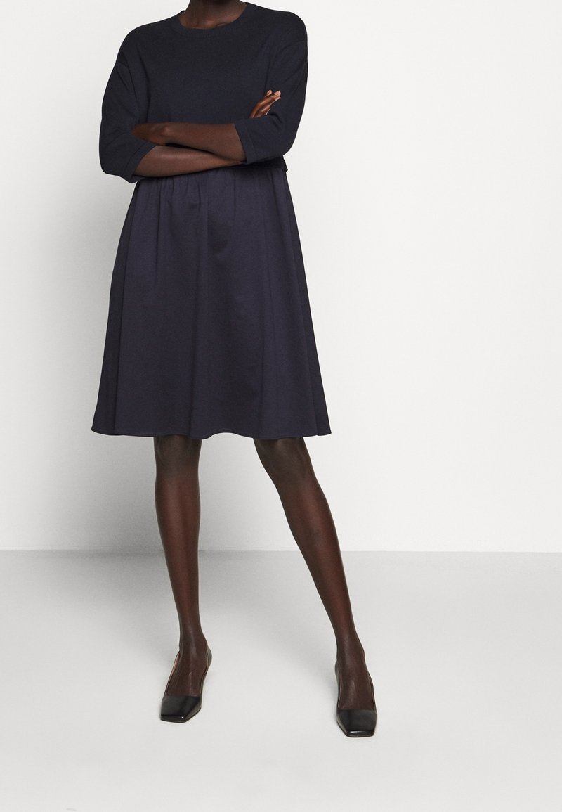 Personne à la peau noire debout, bras croisés, vêtue d'une robe bleu marine foncé jusqu'aux genoux et de chaussures à talons noires sur un fond uni.