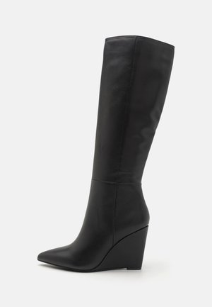 Botte compensée noire montant au genou avec bout pointu et finition en cuir lisse, vue de profil sur fond blanc.