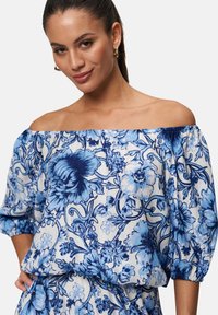 Blouse à épaules dénudées en tissu léger avec une base blanche, ornée de motifs floraux bleus, de manches bouffantes et d'un col élastique.