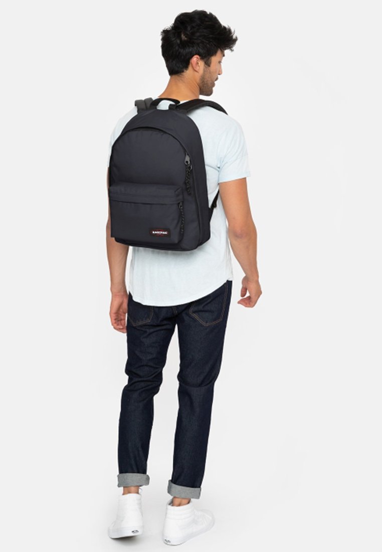 Un sac à dos en tissu noir avec des bretelles réglables, une poche avant et un patch de logo. Le porteur porte des jeans retroussés et des baskets blanches.