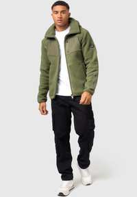 Olivengrüne Fleecejacke mit Reißverschluss vorne, Kapuze und mehreren Taschen; kombiniert mit schwarzen Cargo-Hosen und weißen Sneakern.