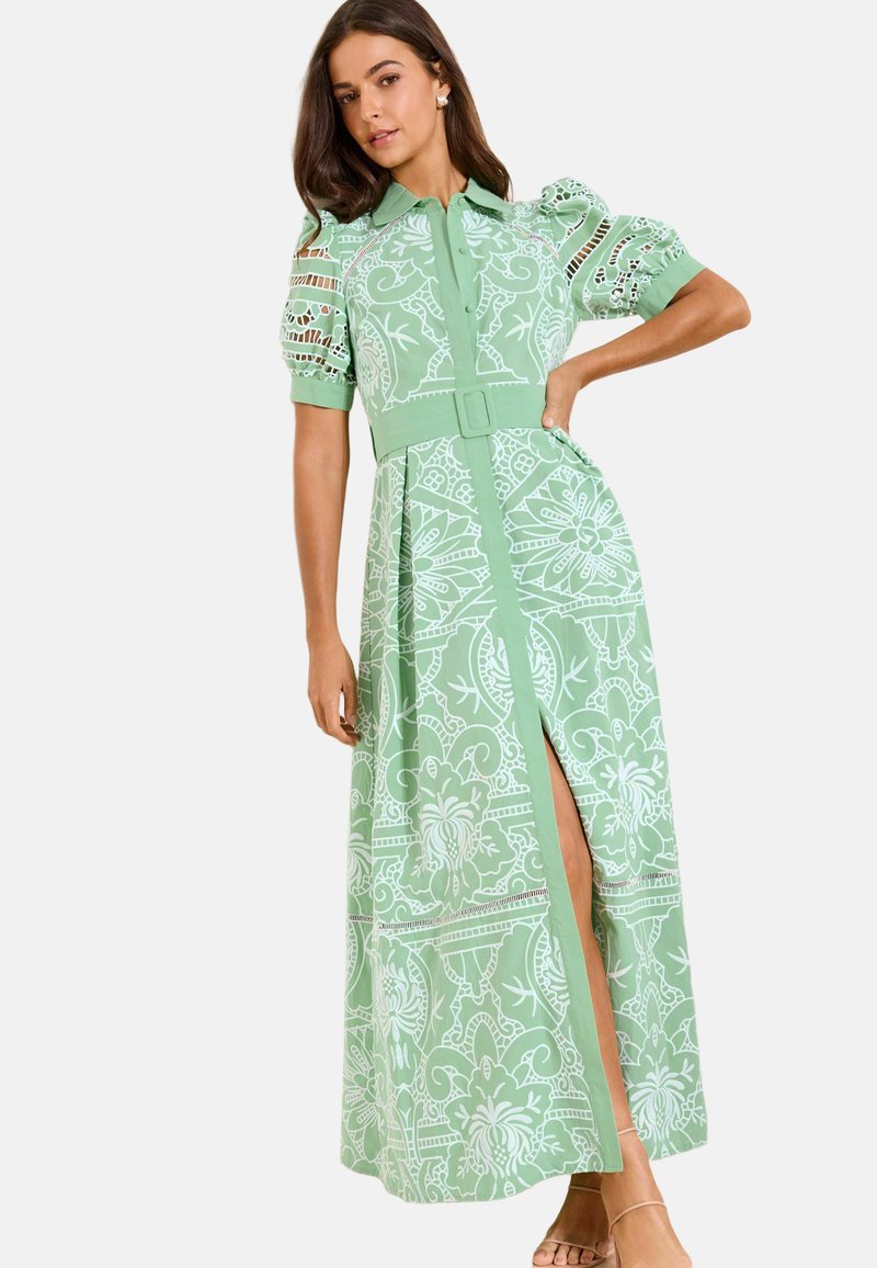 Sieviete tērpusies zaļā gara kleitā ar rakstu, pūkainām piedurknēm, jostu un priekšējo šķēlumu, pozējot ar vienu roku uz gurna uz vienkārša fona.