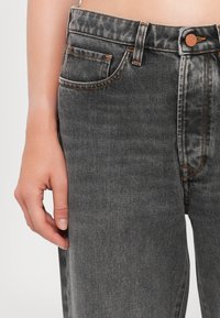 Gros plan sur la main gauche d’une personne reposant à côté d’un jean en denim noir taille haute avec des coutures visibles et un détail de bouton.