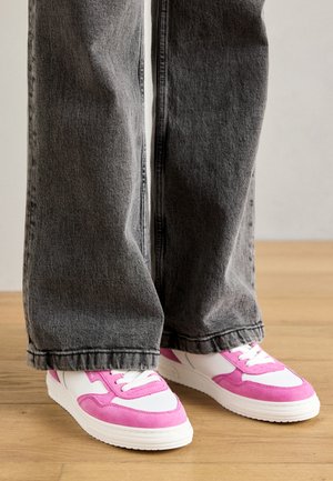 Des chaussures de sport blanches et roses avec des accents en daim, associées à un jean large gris foncé, posées sur un sol en bois.