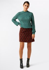 Teal strikket sweater med høj hals, parret med en tætsiddende rød nederdel med blomstermønster. Sorte ankelstøvler med en glansfuld finish.