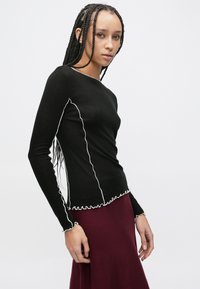 Bluza neagră cu mâneci lungi, cusături albe cu volane, o croială mulată, decolteu rotund și țesătură texturată. Asortată cu o fustă burgundy.