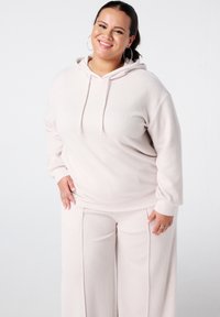 Lichtroze geribde hoodie met kangoeroezak en trekkoord, gecombineerd met bijpassende wijde ribbroek. Model glimlacht en draagt houten oorbellen.