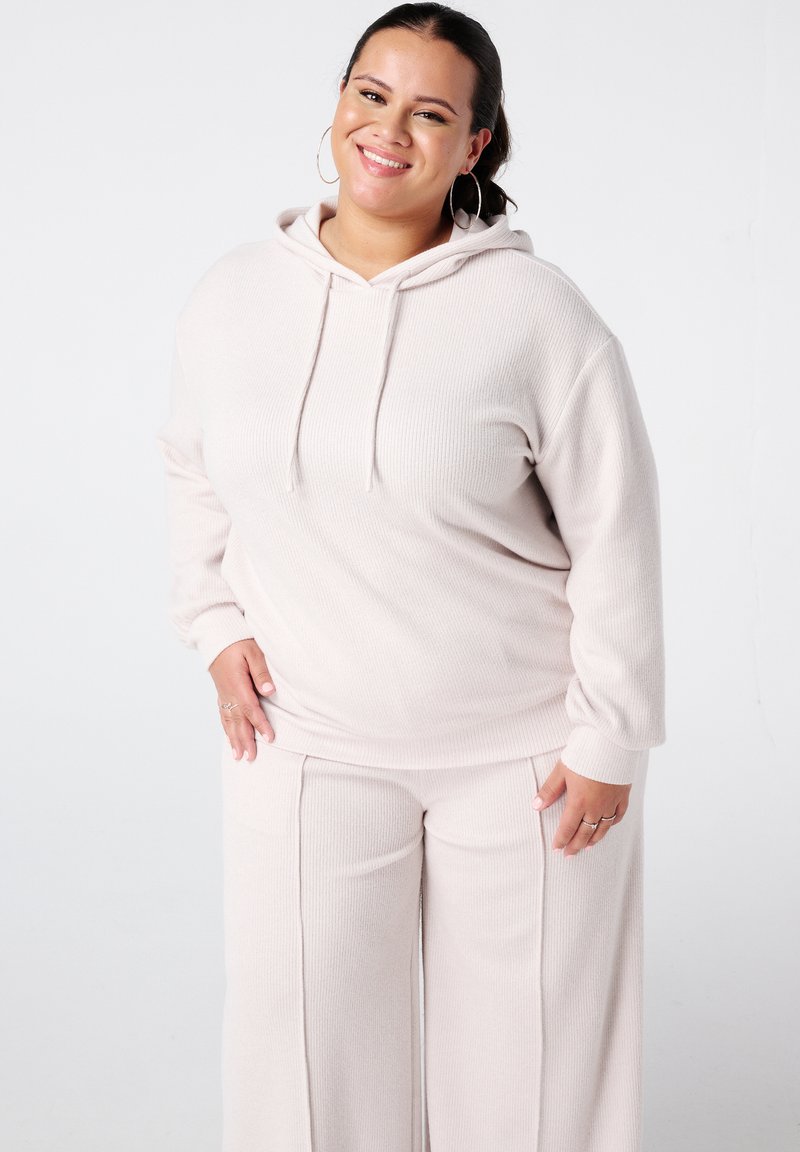 Lichtroze geribde hoodie met kangoeroezak en trekkoord, gecombineerd met bijpassende wijde ribbroek. Model glimlacht en draagt houten oorbellen.